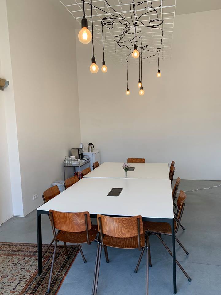 Kleine zaal met 11 stoelen rond een centrale tafel en koelkast in de hoek